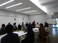 室内にテーブルが置かれ、グループに分かれて参加者が着席している。写真の右手に講師が立って話をしており、参加者は講師の方を向いている。