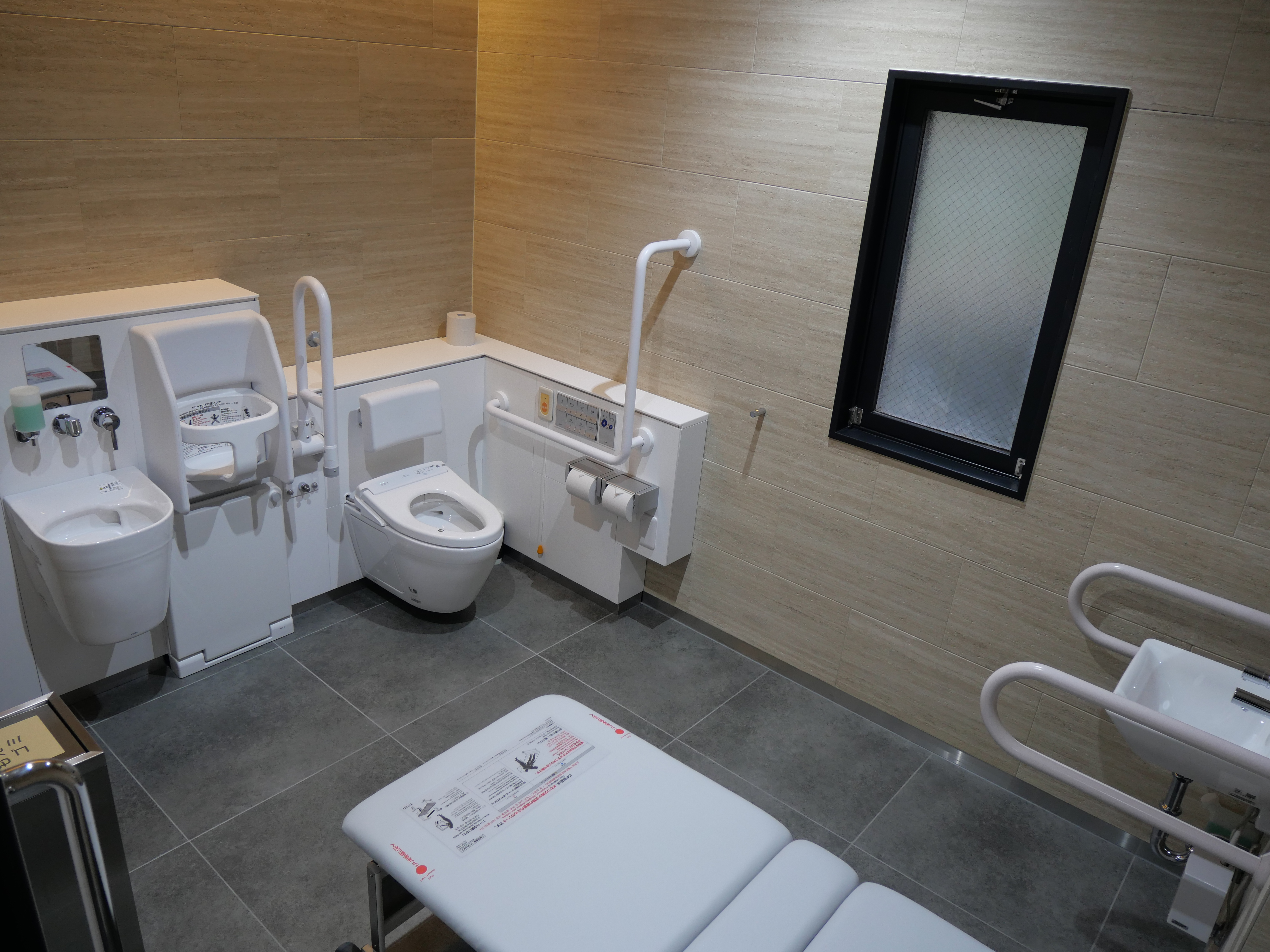 美術情報室前の多機能トイレ。入って左手にオストメイト対応のシンク、鏡、着替用シートがあり、ドアの近くに折り畳み式のユニバーサルシート（介助用シート）がある。