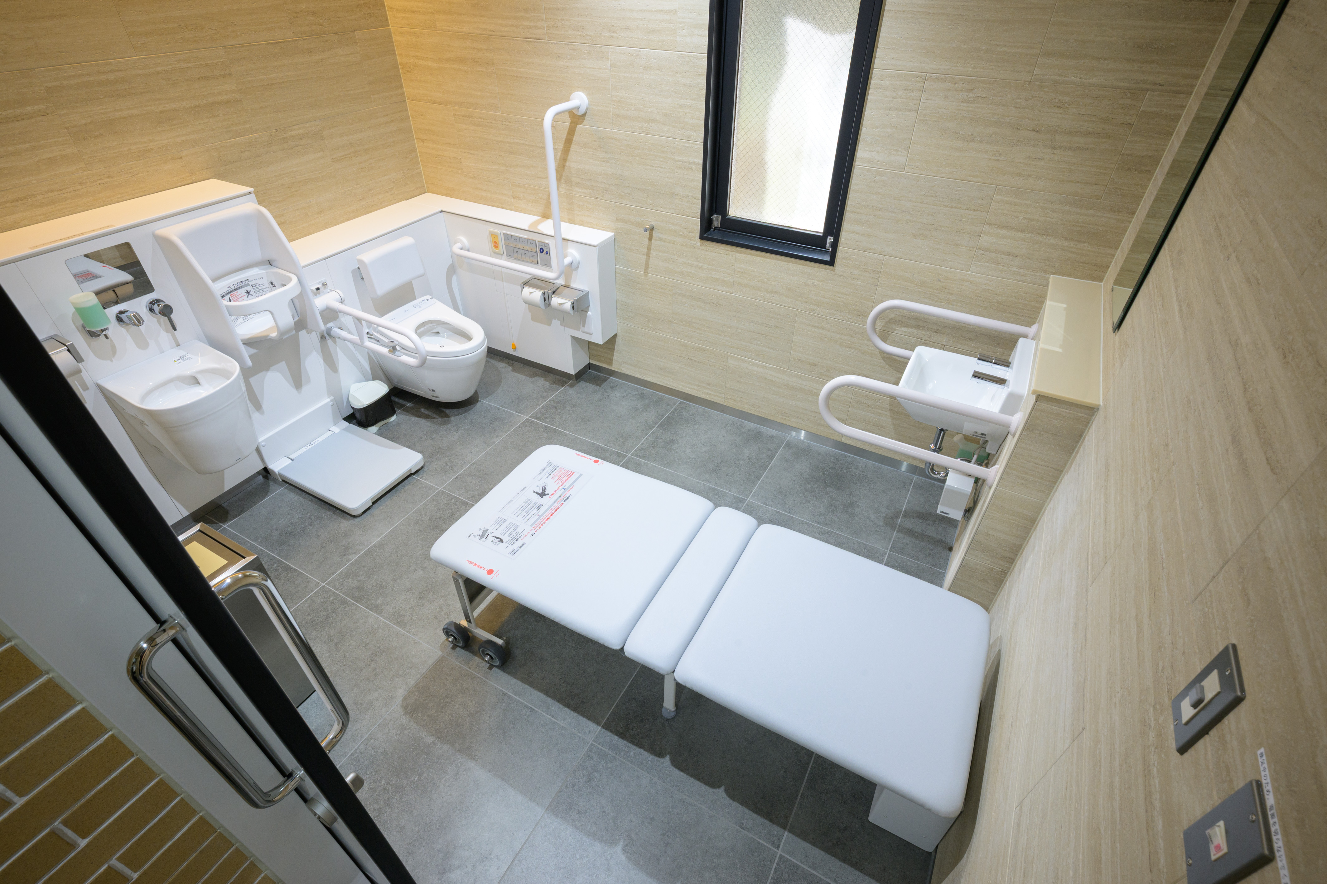 美術情報室前の多機能トイレ。入って左手にオストメイト対応のシンク、鏡、着替用シートがあり、ドアの近くに折り畳み式のユニバーサルシート(介助用シート)がある。