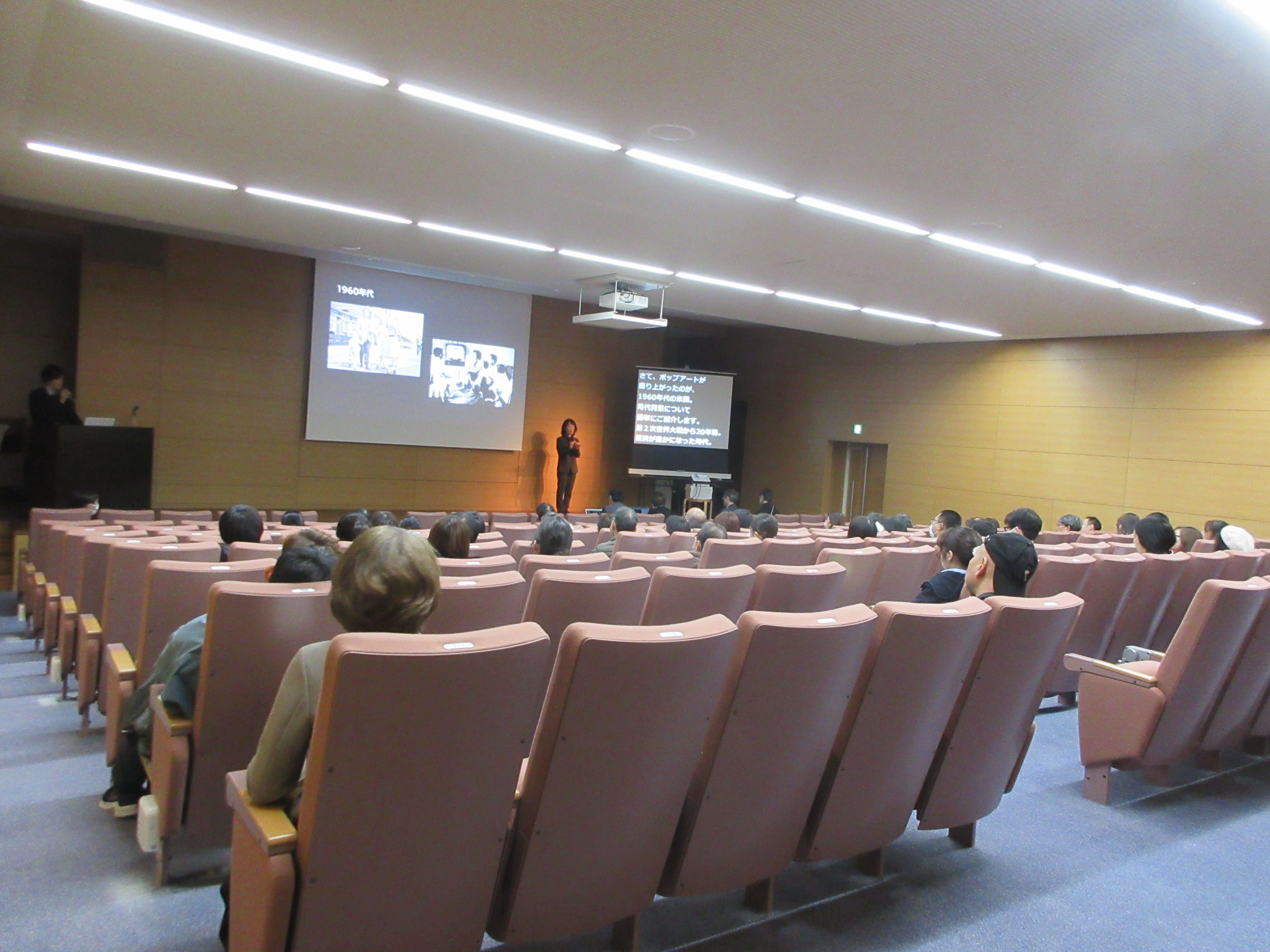 講演会の様子。中央のスクリーンに画像が映され、右手に手話通訳者が立つ。その右に文字が映ったスクリーンがある